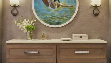 Un baño con tocador de madera, un espejo redondo, dos luces fijadas a la pared, un jarrón con flores y un cuadro de un nadador.