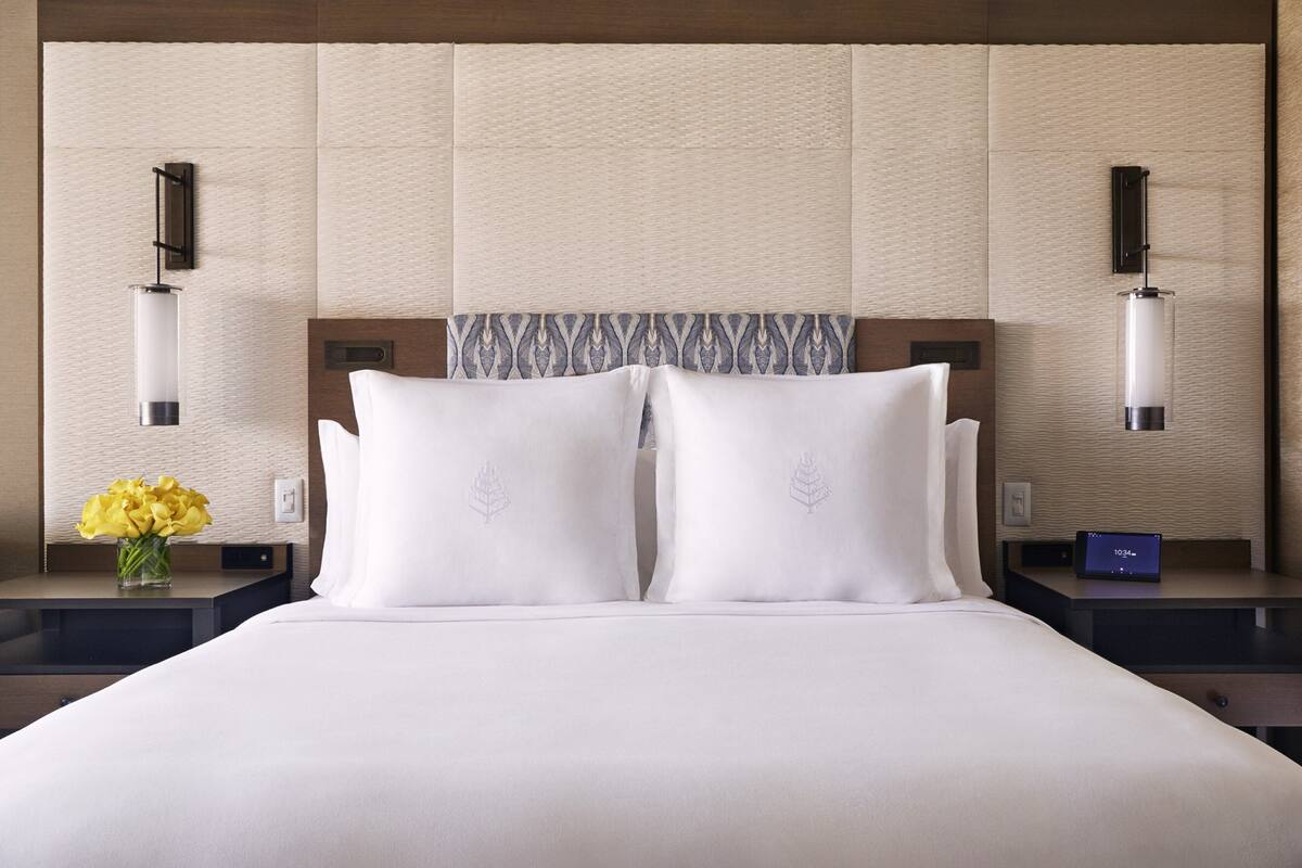 Une chambre d’hôtel avec un grand lit, deux tables de chevet, une horloge numérique et un vase rempli de fleurs jaunes.
