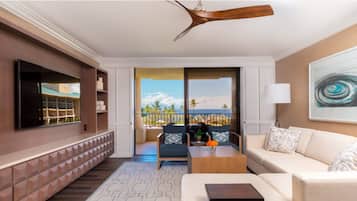 A modern living room with a flat-screen TV, a ceiling fan, a sofa, a coffee table, and a balcony with a view of the ocean.
