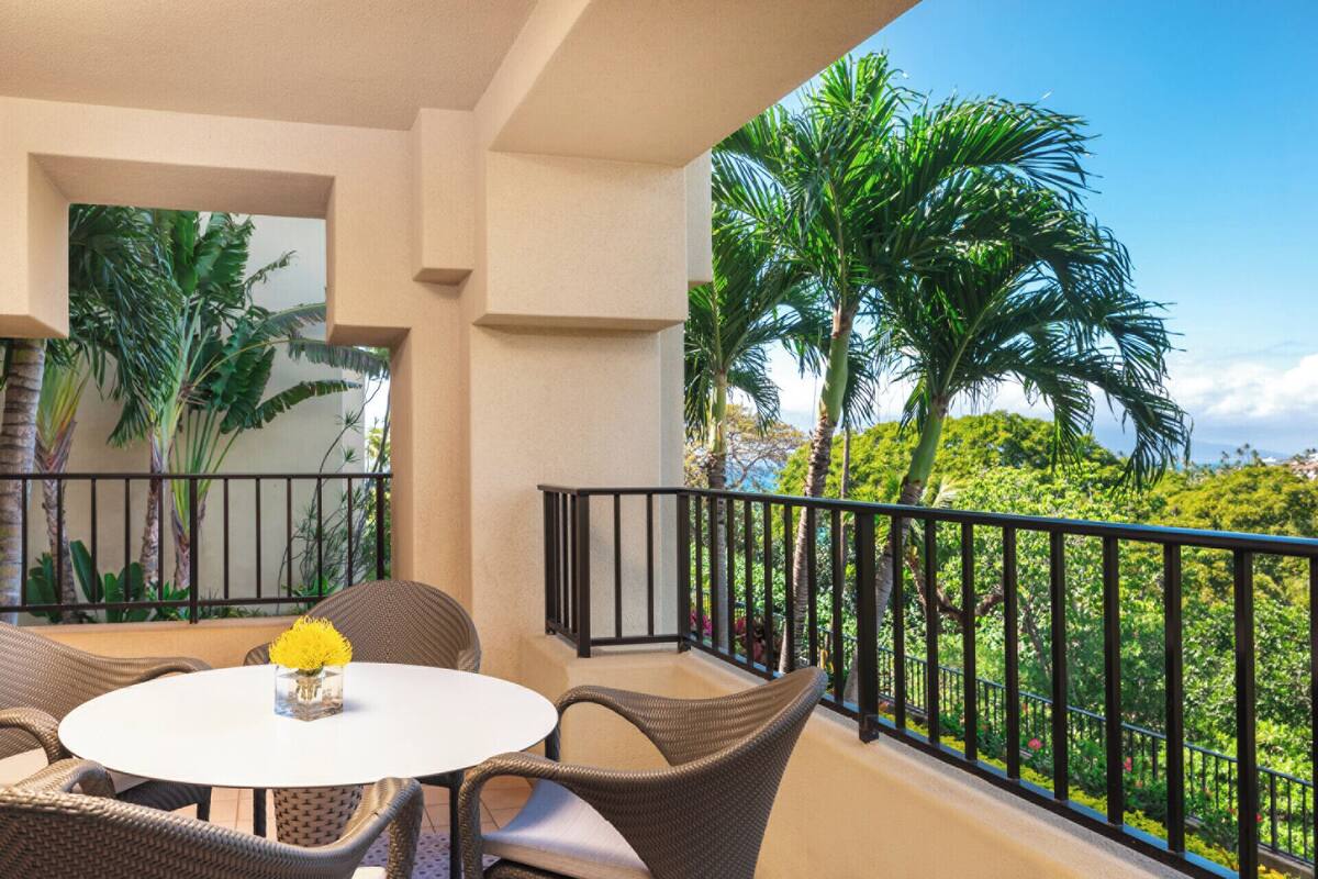 Un balcon agrémenté de meubles en osier et d’un vase rempli de fleurs jaunes, offrant une vue sur un paysage tropical avec des palmiers et un ciel dégagé.