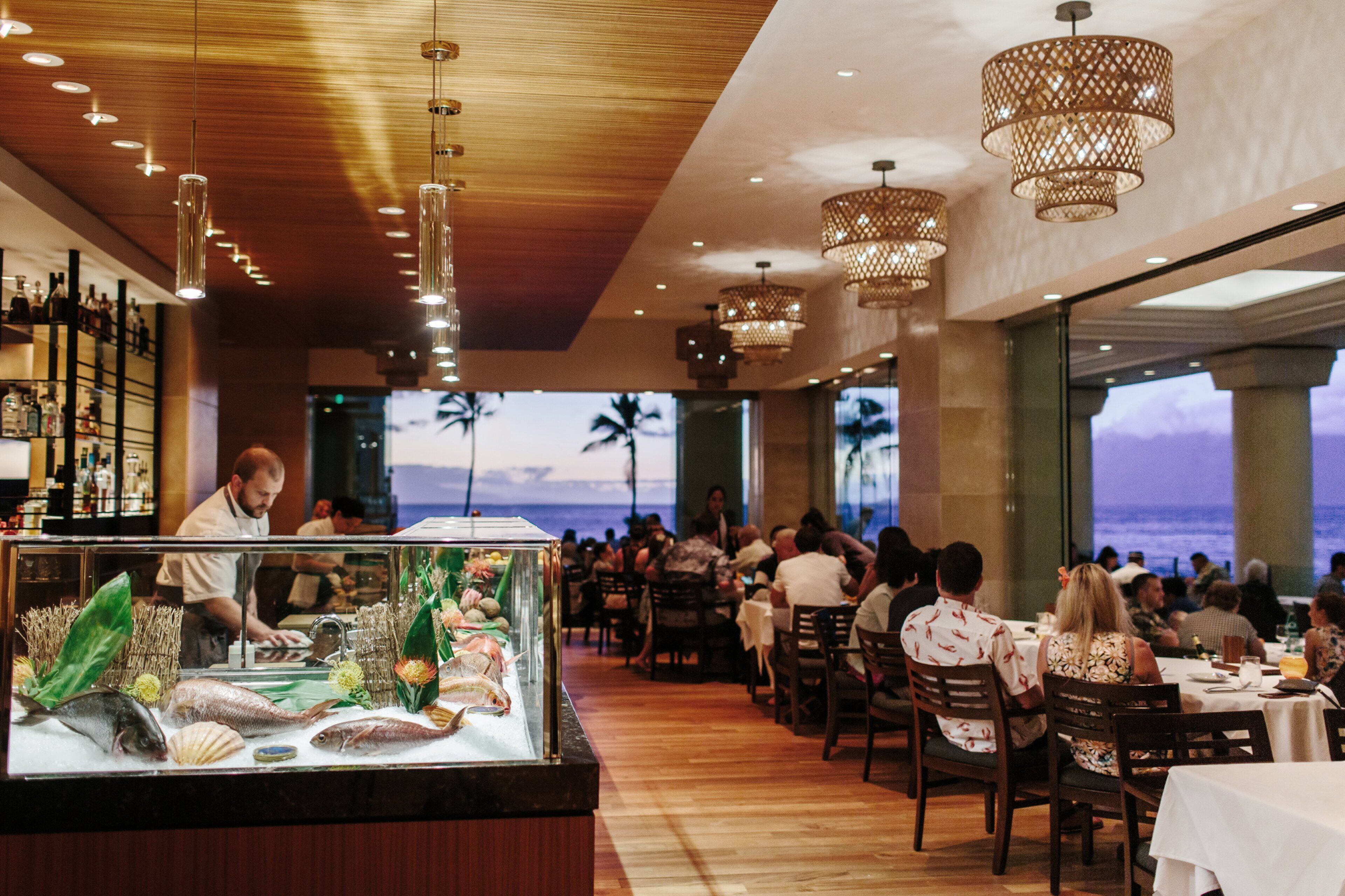 Een restaurant met uitzicht op de oceaan, een houten plafond en hangende kroonluchters.