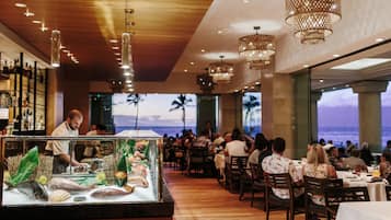 A restaurant with a view of the ocean, wooden ceiling, and hanging chandeliers.