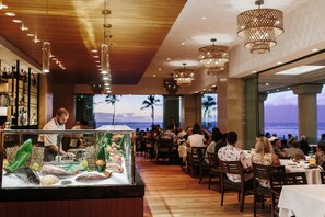 A restaurant with a view of the ocean, wooden ceiling, and hanging chandeliers.