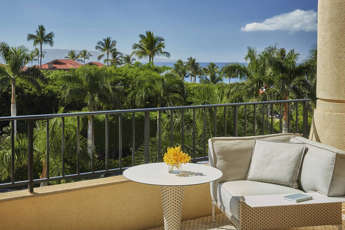 Un balcon avec une table blanche et un canapé beige, donnant sur un paysage tropical avec des palmiers et une vue sur l’océan.