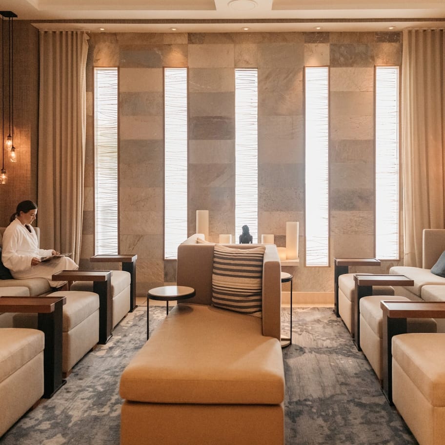 A modern hotel room with a central seating area, multiple sofas, and a large window with curtains.