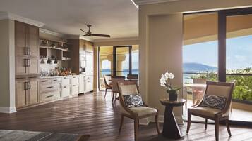 A spacious living area with a kitchen, two chairs with cushions, a ceiling fan, and a view of the ocean.