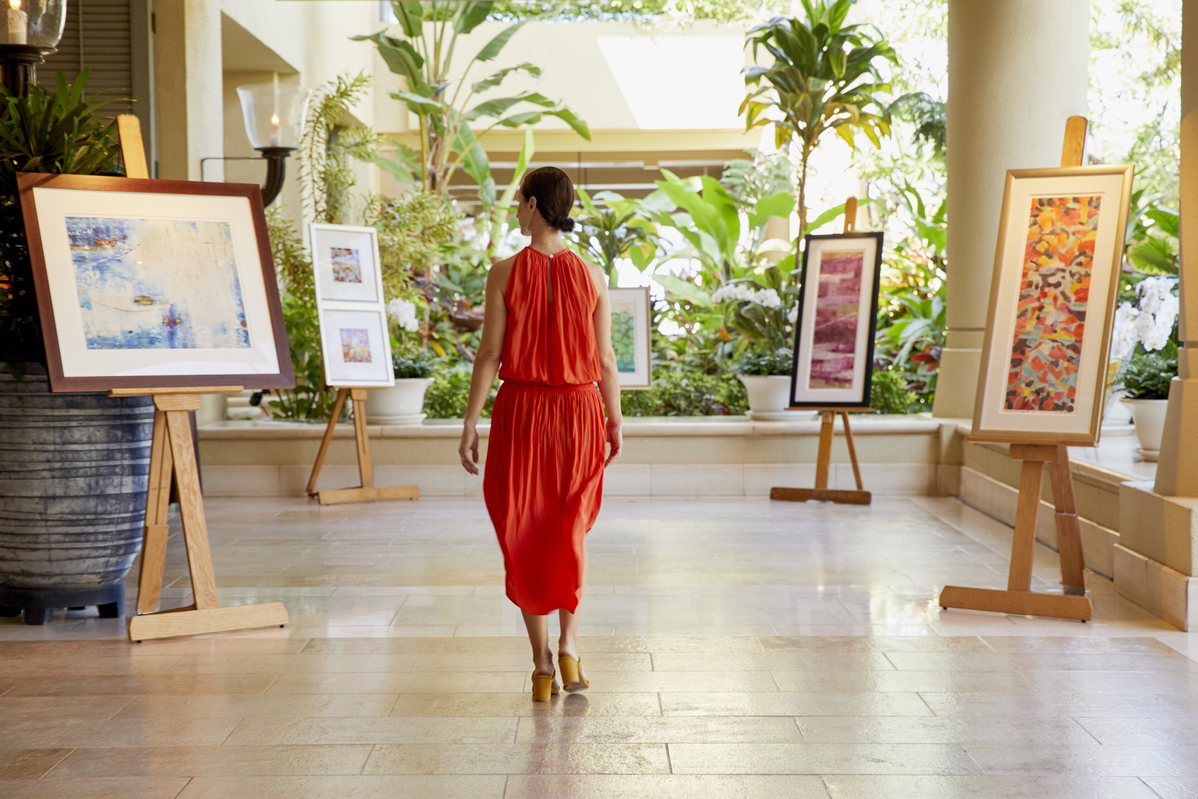 A woman in a red dress walking through a lobby with art on easels and potted plants.