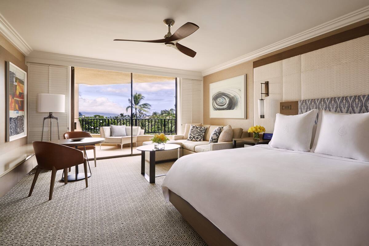 Une chambre d’hôtel avec un grand lit, un canapé, une table basse, un fauteuil et un balcon donnant sur des palmiers.