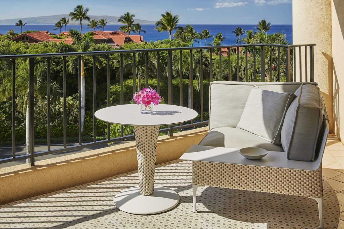 Un balcon avec une table ronde et un canapé, un vase rempli de fleurs roses, et une vue sur l’océan et les palmiers.