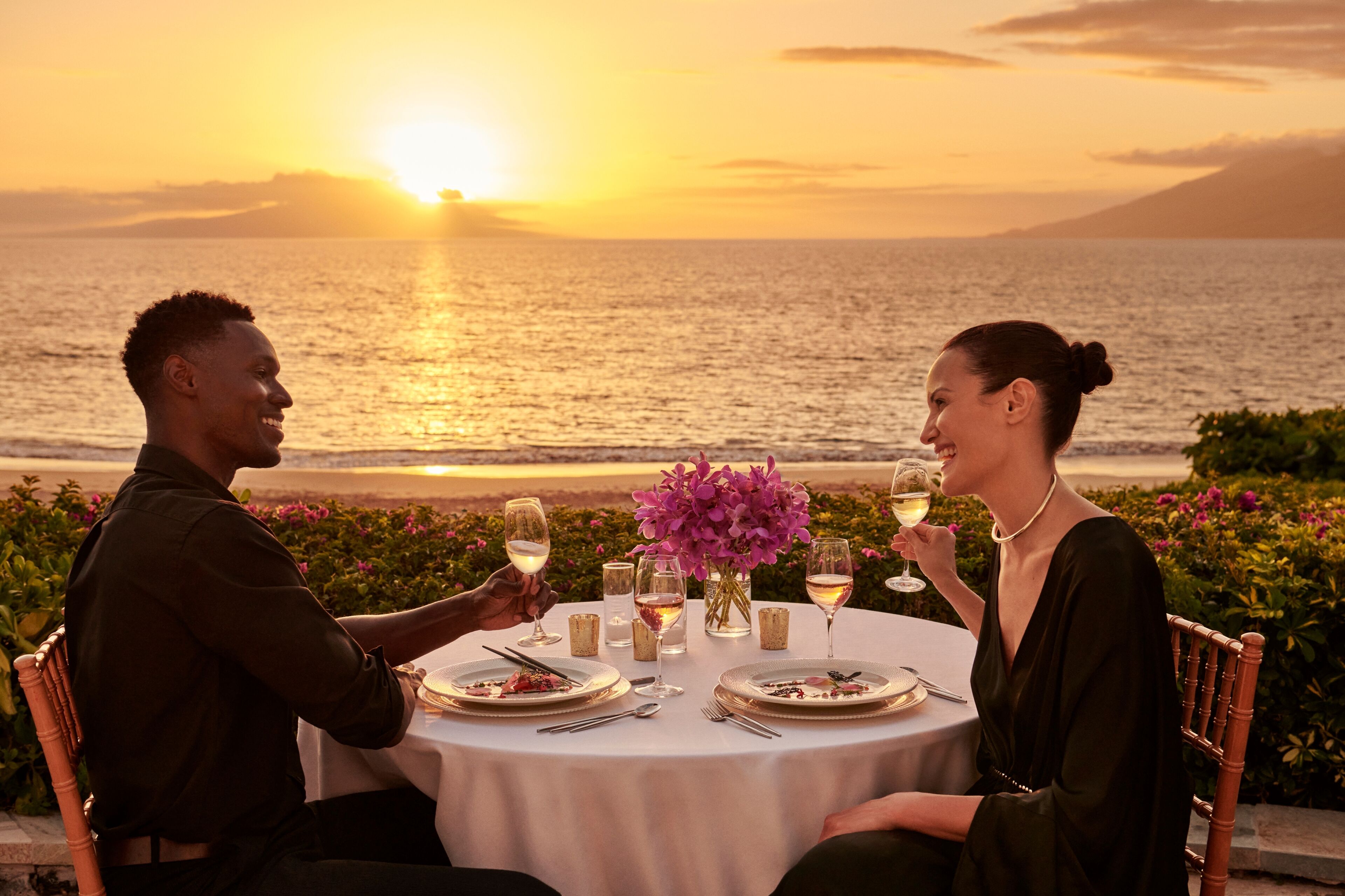 A couple dining at a table with a view of the sunset over the ocean.