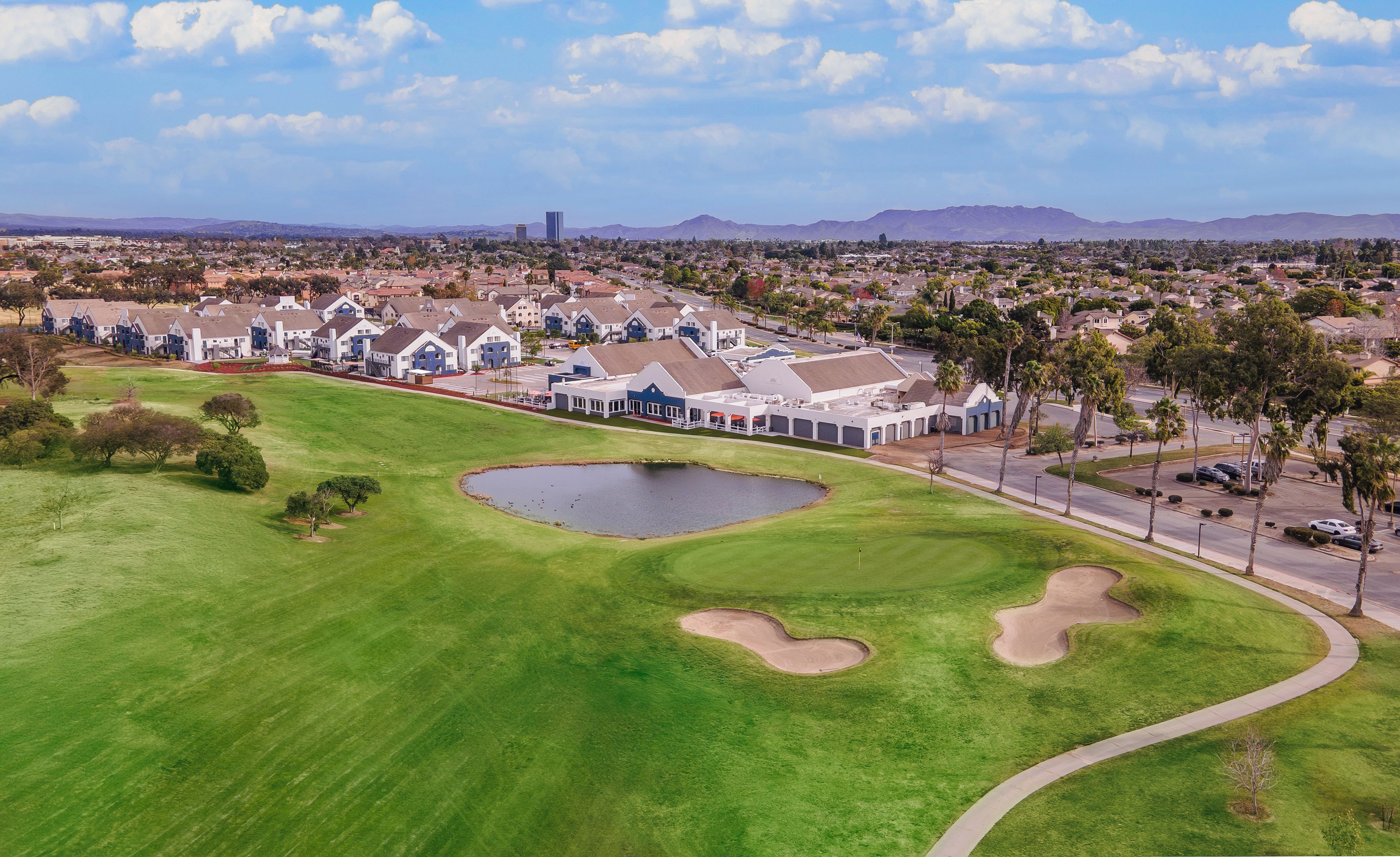 Ein Golfplatz mit Teich, Clubhaus und Wohnhäusern im Hintergrund.