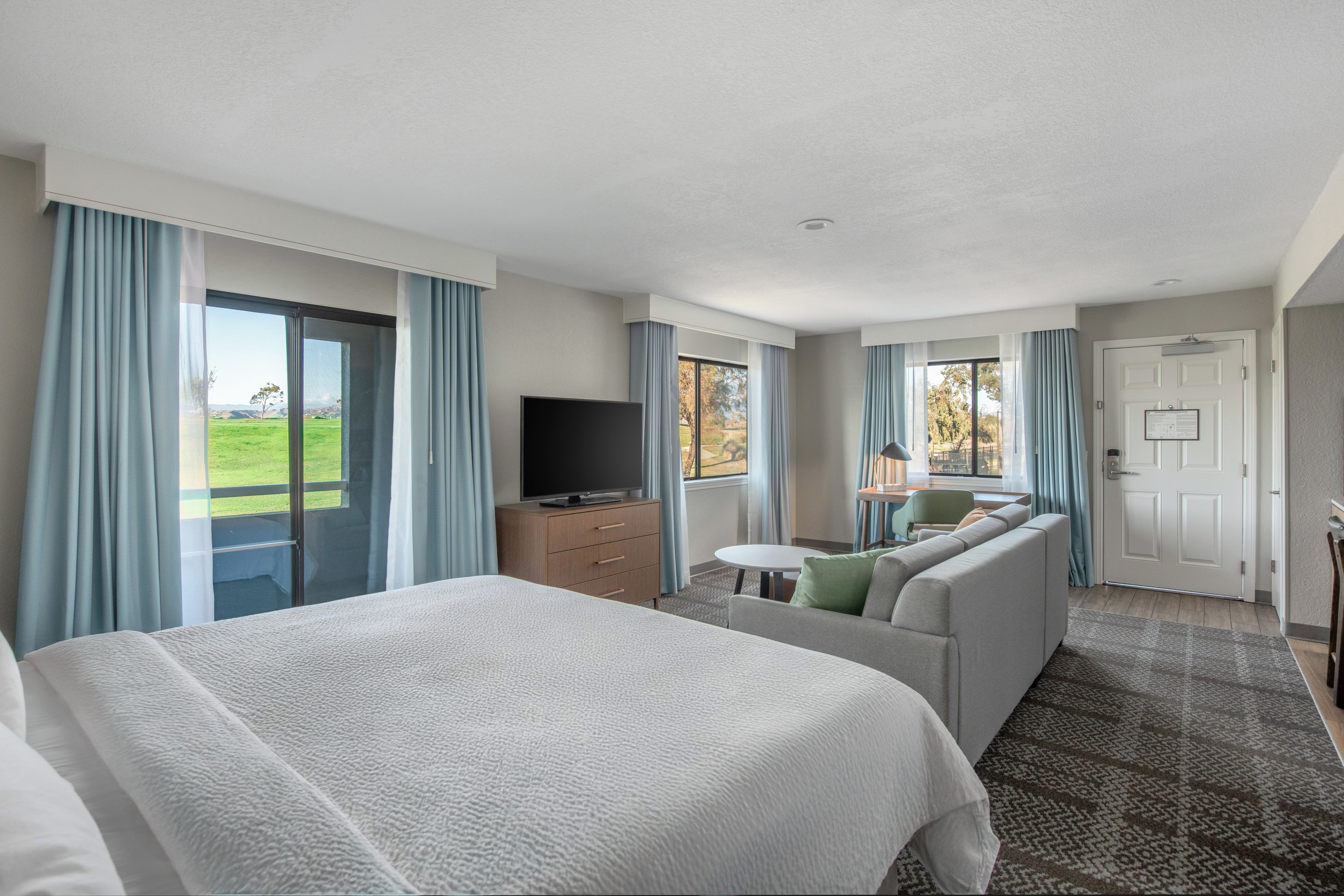 A hotel room with a large bed, a sofa, a television, and a view of the outdoors.