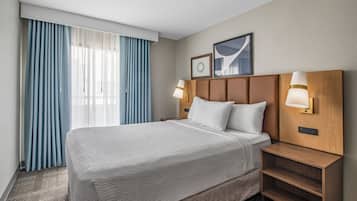 A hotel room with a large bed, two bedside tables, a headboard with wall-mounted lights, a window with blue curtains, and a framed picture on the wall.