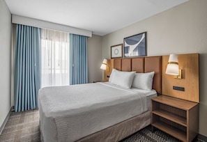 A hotel room with a large bed, two bedside tables, a headboard with wall-mounted lights, a window with blue curtains, and a framed picture on the wall. - Staybridge Suites Oxnard River Ridge by IHG (Oxnard)