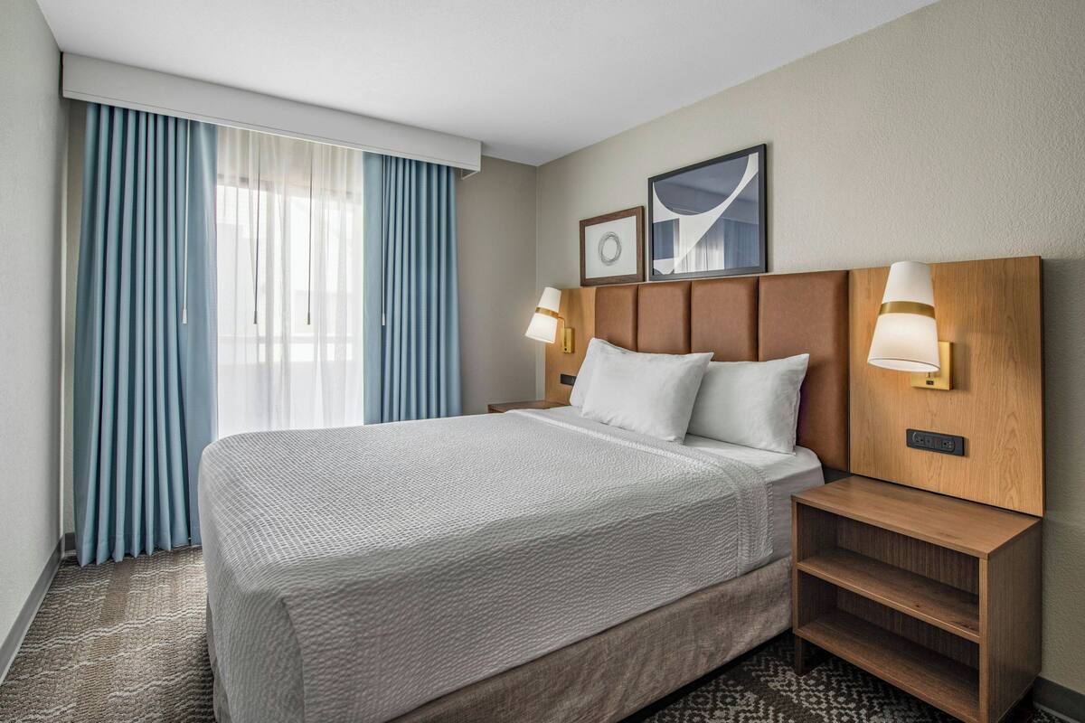 A hotel room with a bed, two bedside lamps, a headboard, a nightstand, a carpet, and a window with curtains.