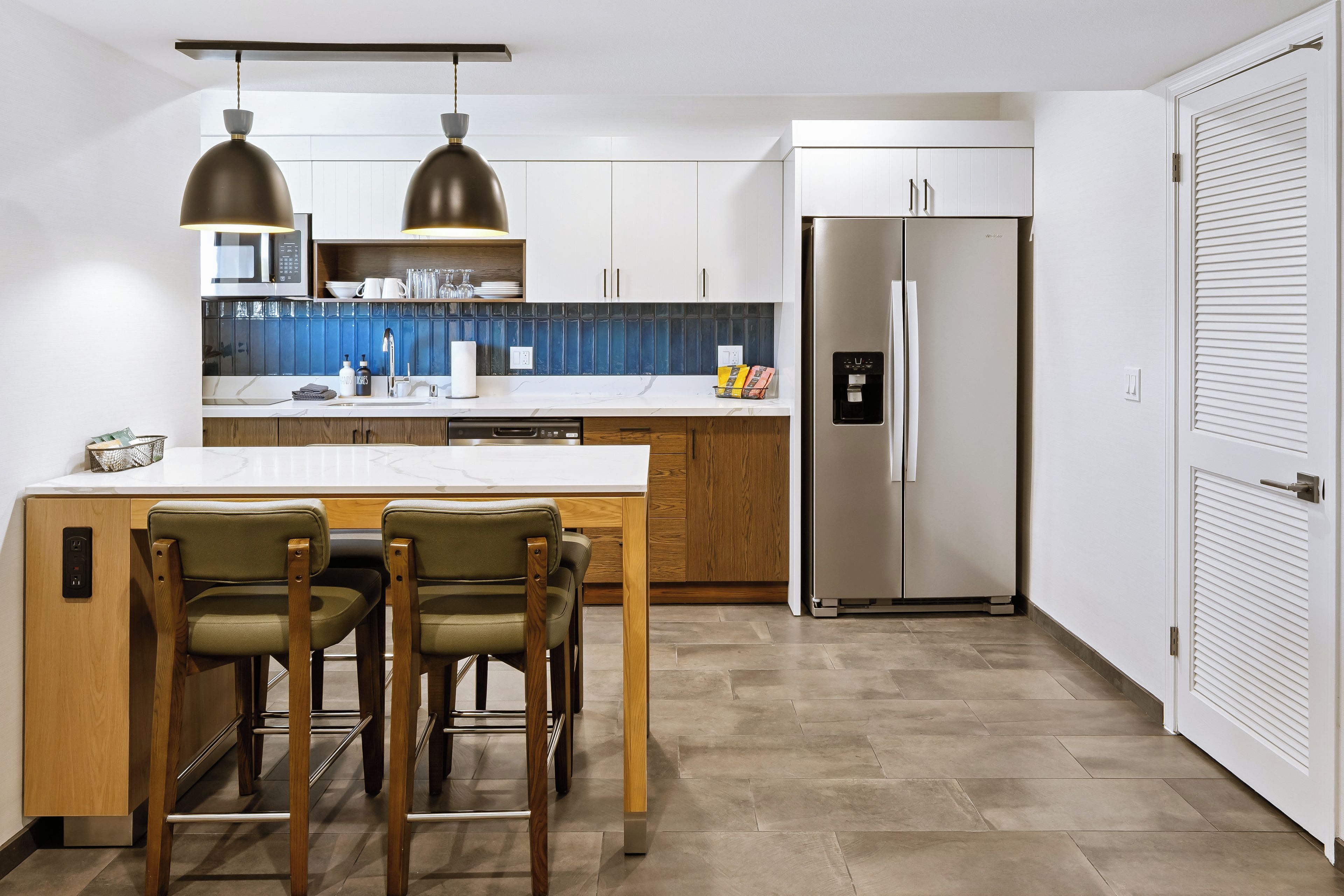 A modern kitchen with a central island, two pendant lights, a built-in microwave, and a stainless steel refrigerator.