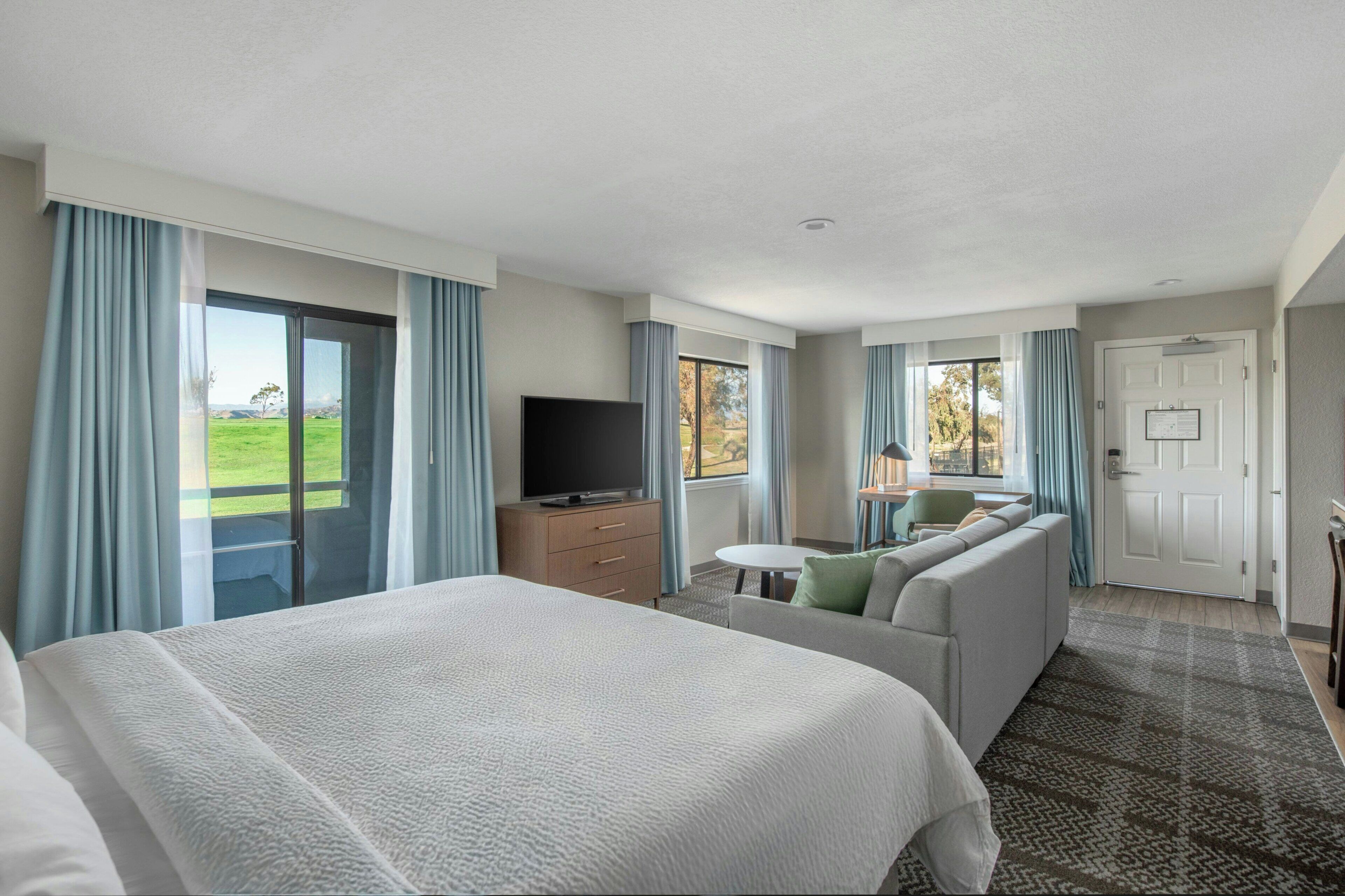 Habitación de hotel con una cama grande, un sofá, un televisor y vistas a un paisaje verde a través de las ventanas.