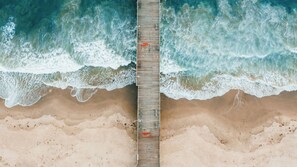 Eine hölzerne Mole, die sich in einen Sandstrand erstreckt, wobei Wellen an beiden Seiten brechen.