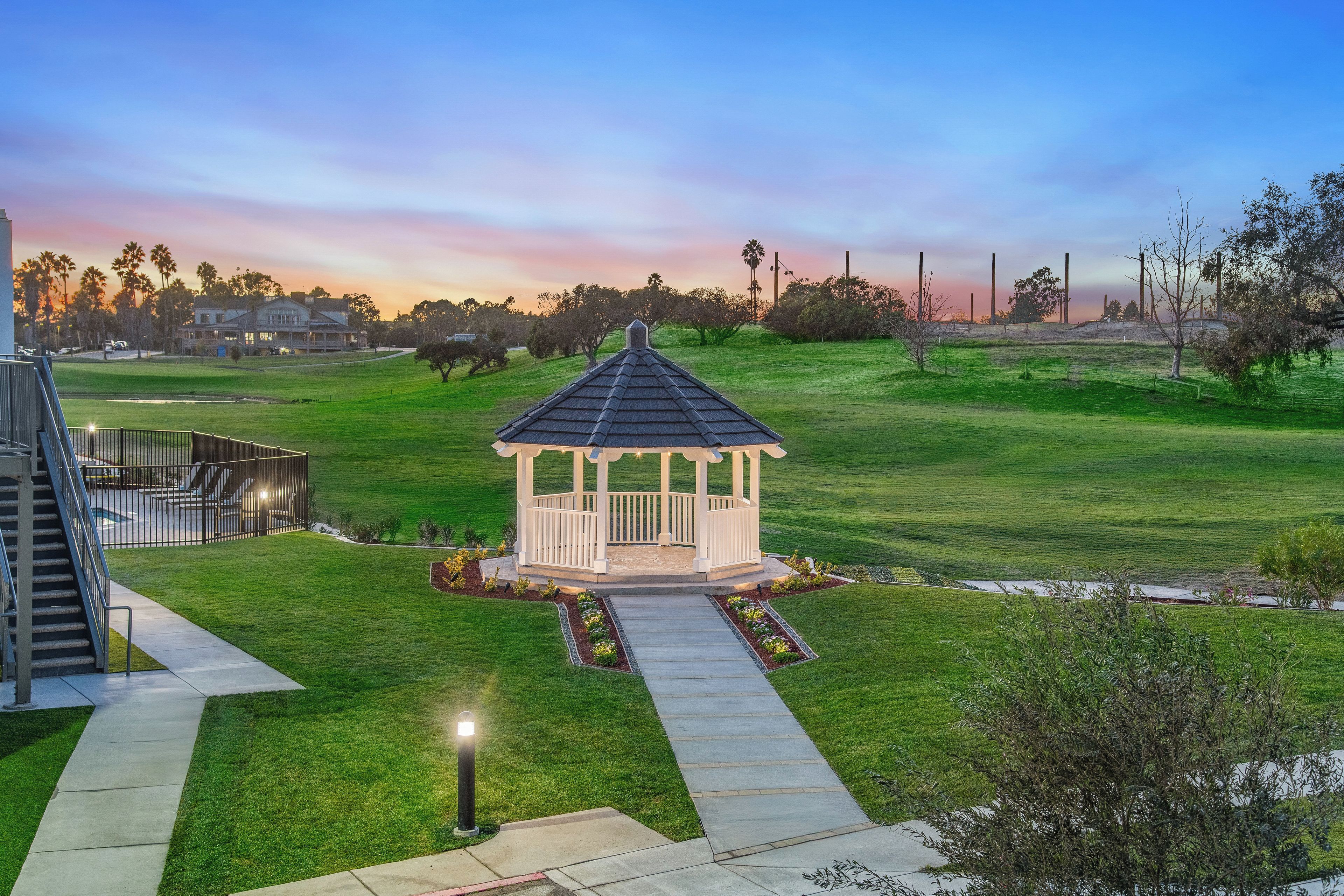 A gazebo in a well-maintained lawn with a pathway leading to it, surrounded by trees and a distant building.