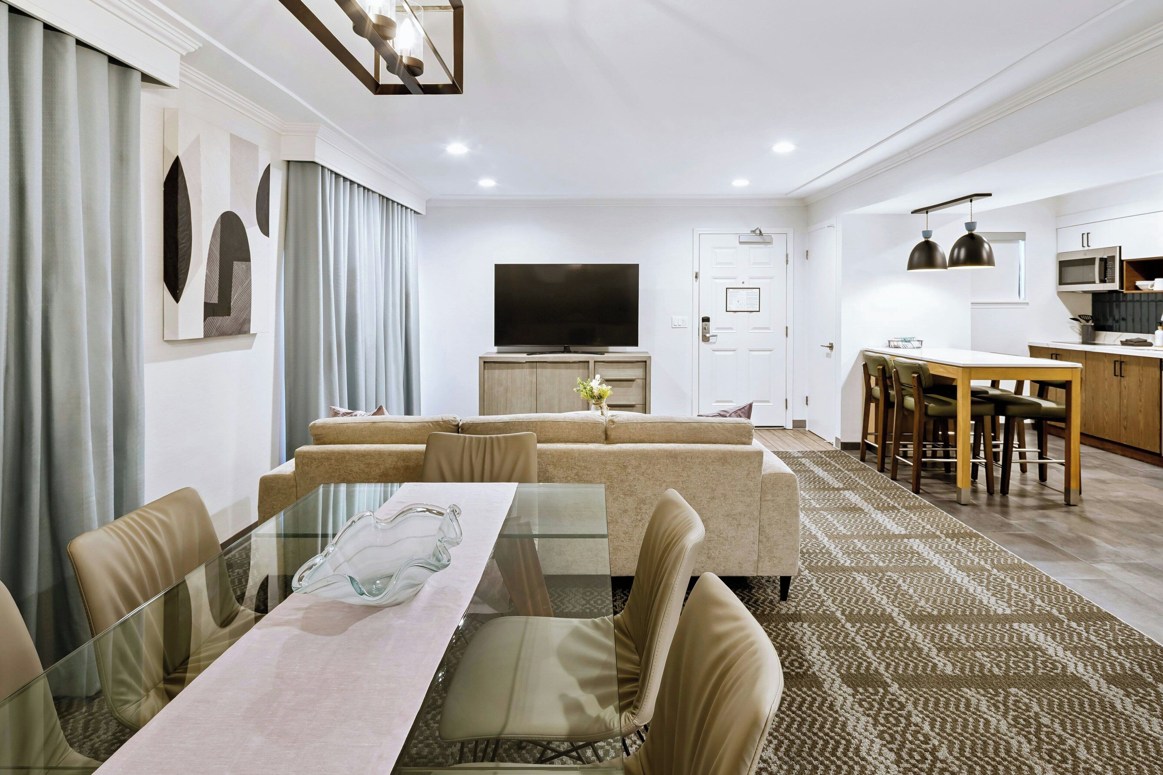 A modern living room with a glass dining table, beige sofas, a flat-screen TV, and a kitchen area with wooden cabinets and pendant lighting.