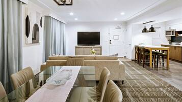 A modern living room with a glass dining table, beige sofas, a flat-screen TV, and a kitchen area with wooden cabinets and pendant lighting.