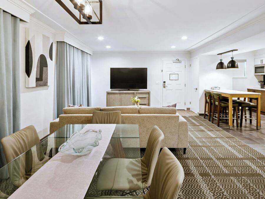 A modern living room with a glass dining table, beige sofas, a flat-screen TV, and a kitchen area with wooden cabinets and pendant lighting.