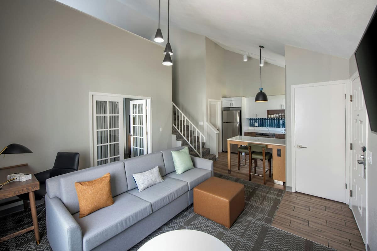 A modern living room with a grey sofa, ottoman, and a staircase leading to a kitchen area.