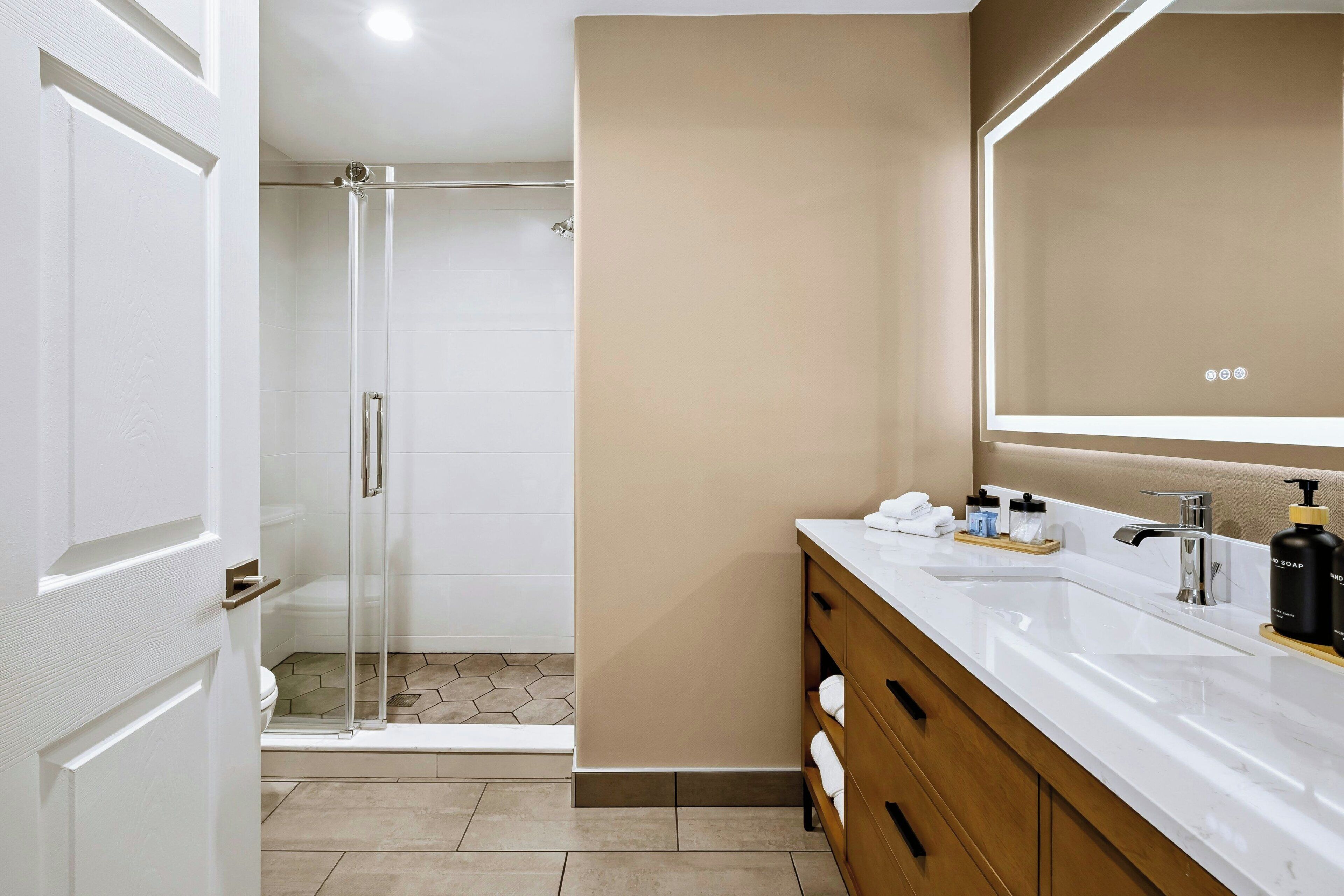 Un baño moderno con cabina de ducha de vidrio, tocador blanco con un espejo grande y pared color beige.