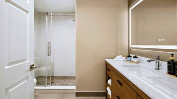 Un baño moderno con cabina de ducha de vidrio, tocador blanco con un espejo grande y pared color beige.