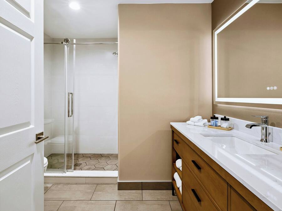 Un baño moderno con cabina de ducha de vidrio, tocador blanco con un espejo grande y pared color beige.