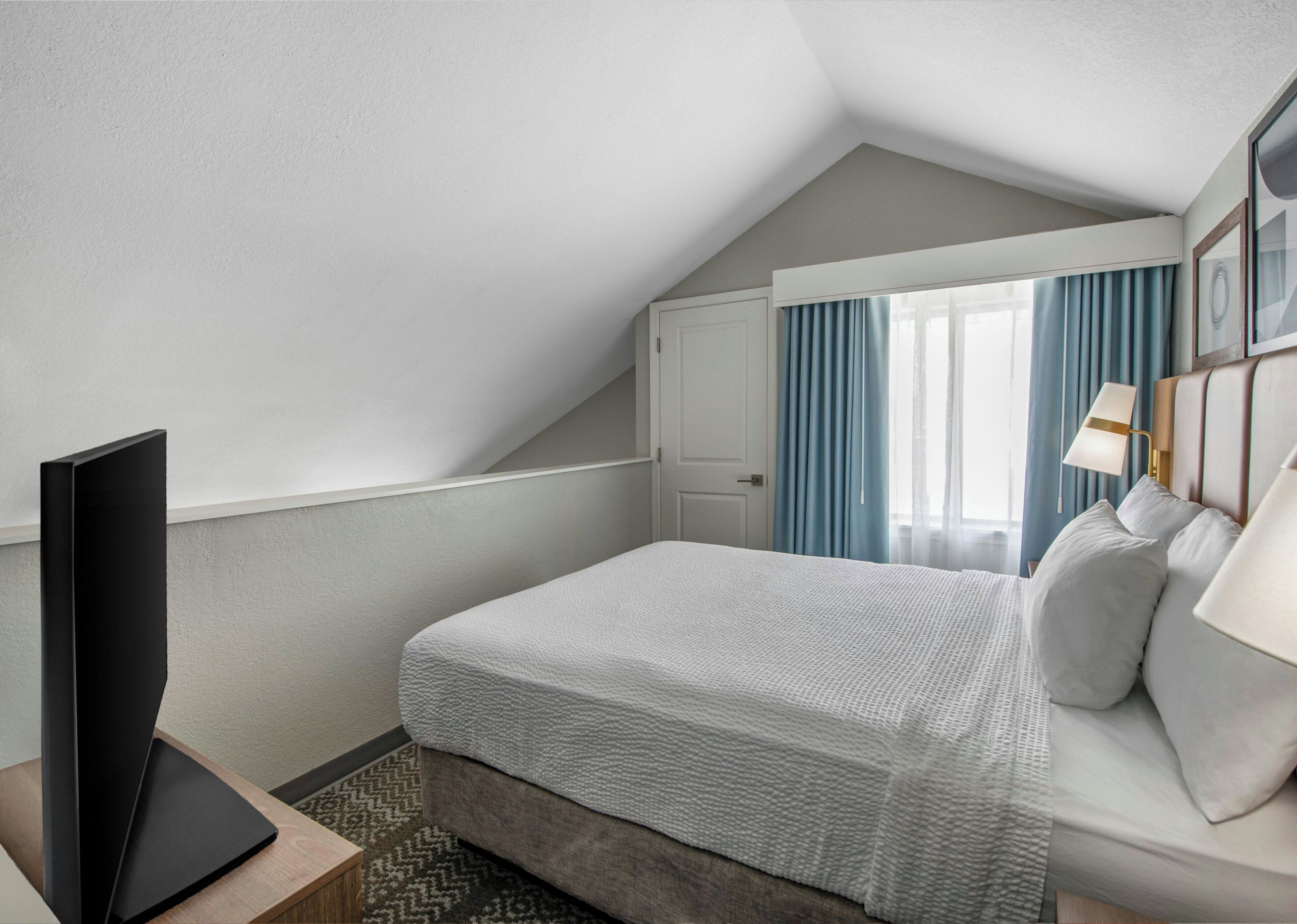 A hotel room with a bed, a television, a desk, and a window with curtains.