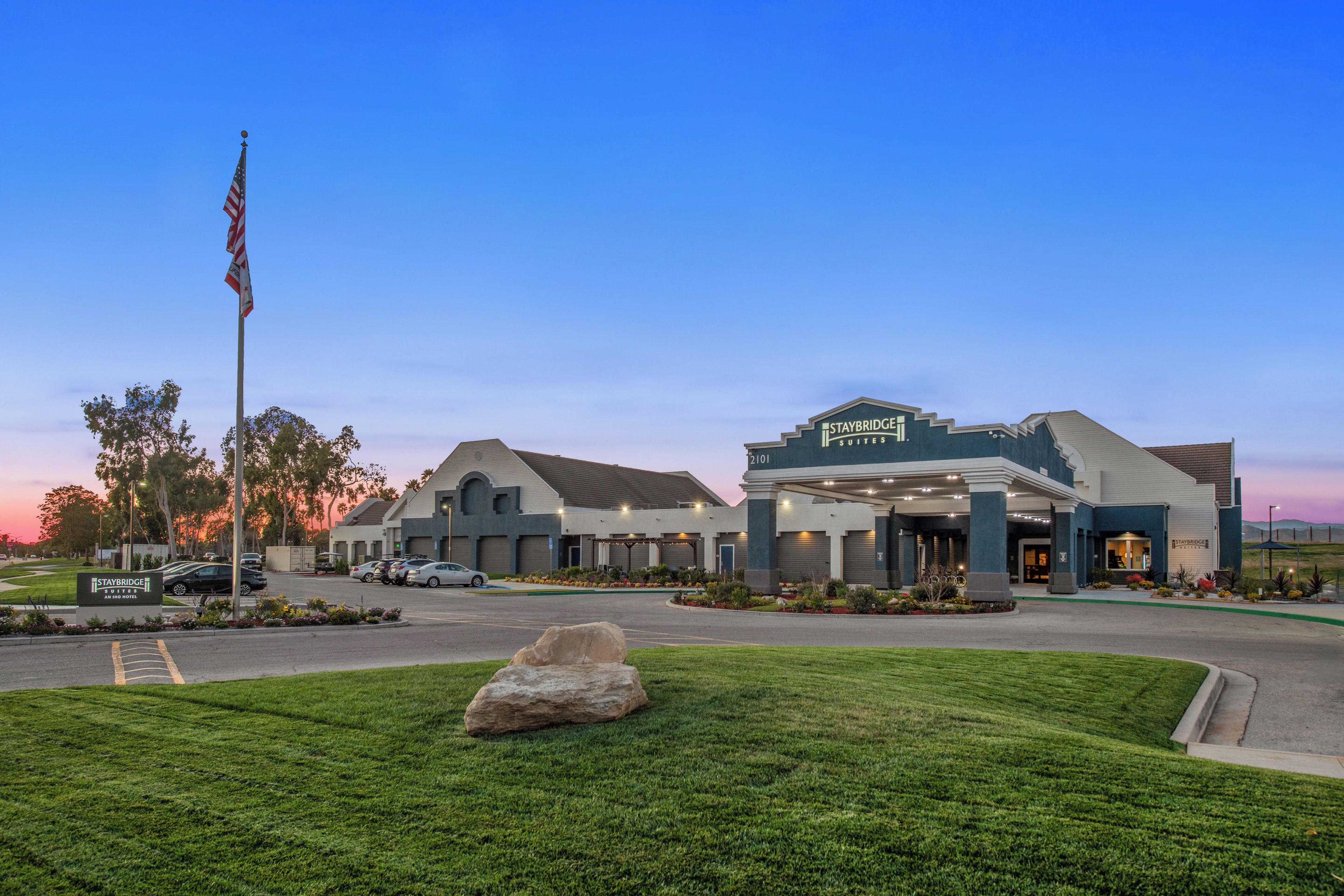 A Staybridge Suites hotel with a large parking lot, a flagpole with the American flag, and well-maintained landscaping.