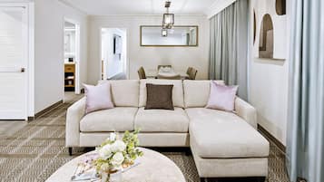 A modern living room with a beige sectional sofa, a round coffee table with a vase of flowers, and a dining area in the background.