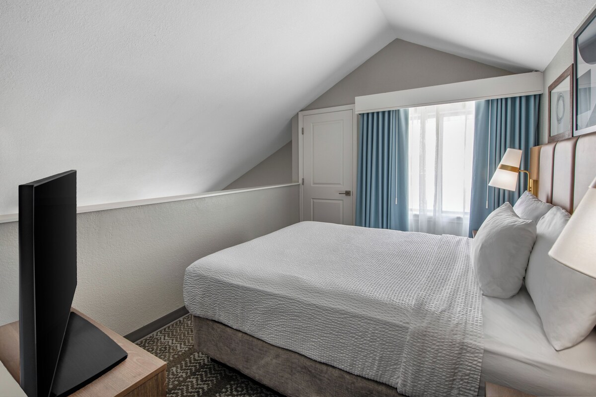 A modern hotel room with a large bed, a flat-screen TV, and a window with curtains.