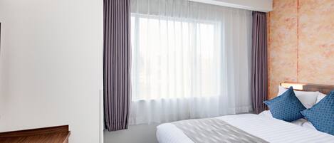A hotel room with a large bed, a wooden desk, and a window with sheer curtains.