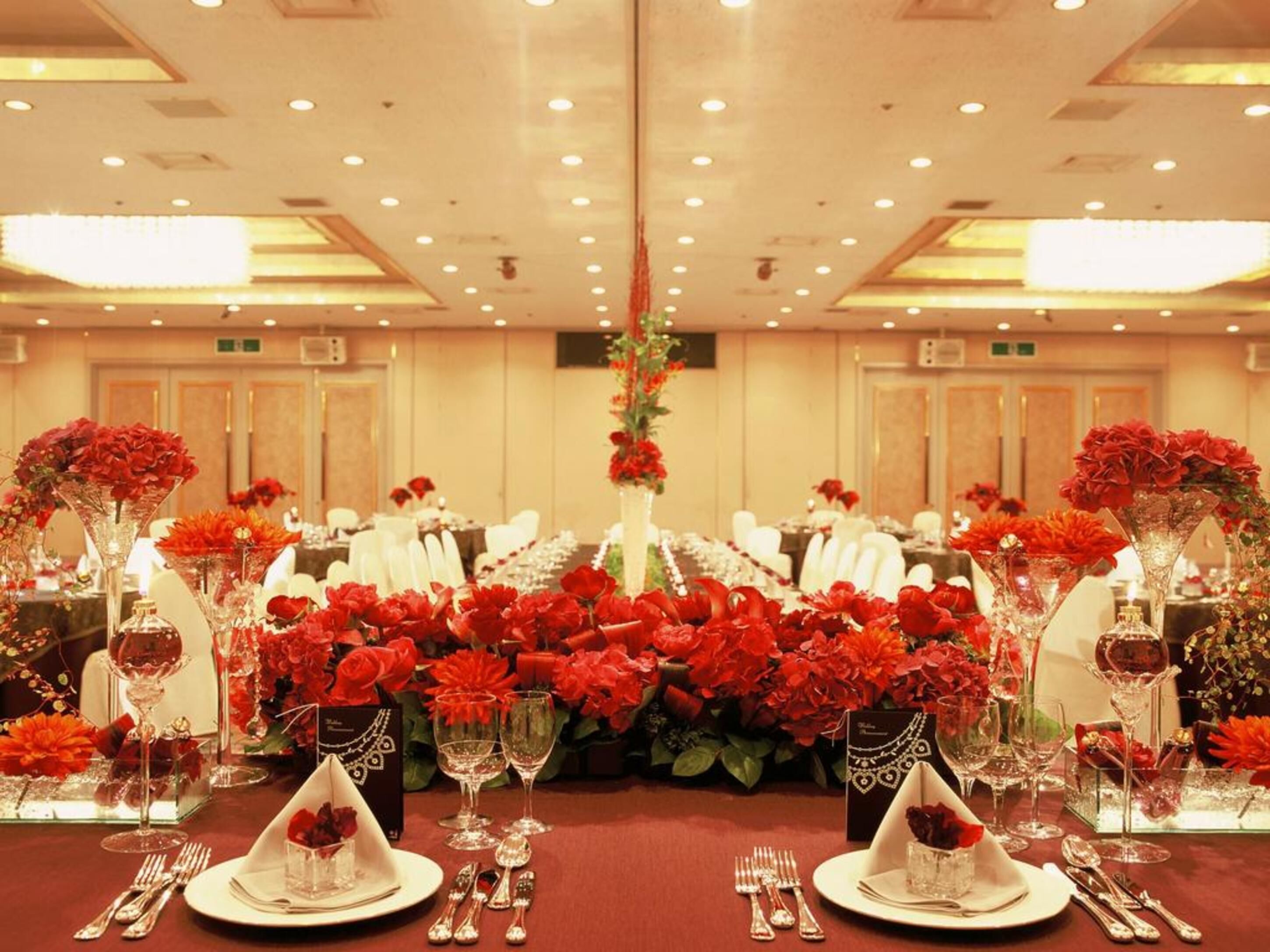 A banquet hall with tables set for a formal event, featuring red floral centerpieces and elegant tableware.
