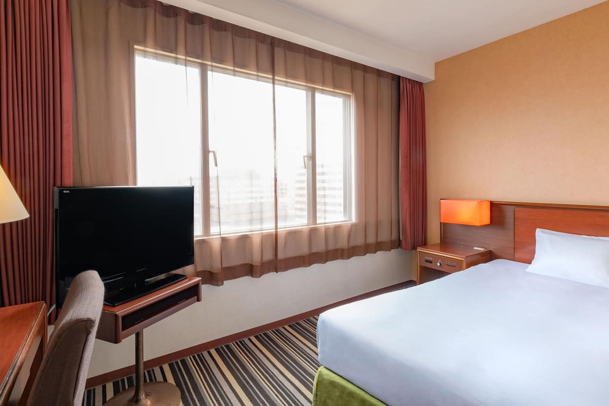 A hotel room with a bed, a TV on a stand, a desk, a chair, a nightstand, and a window with curtains.