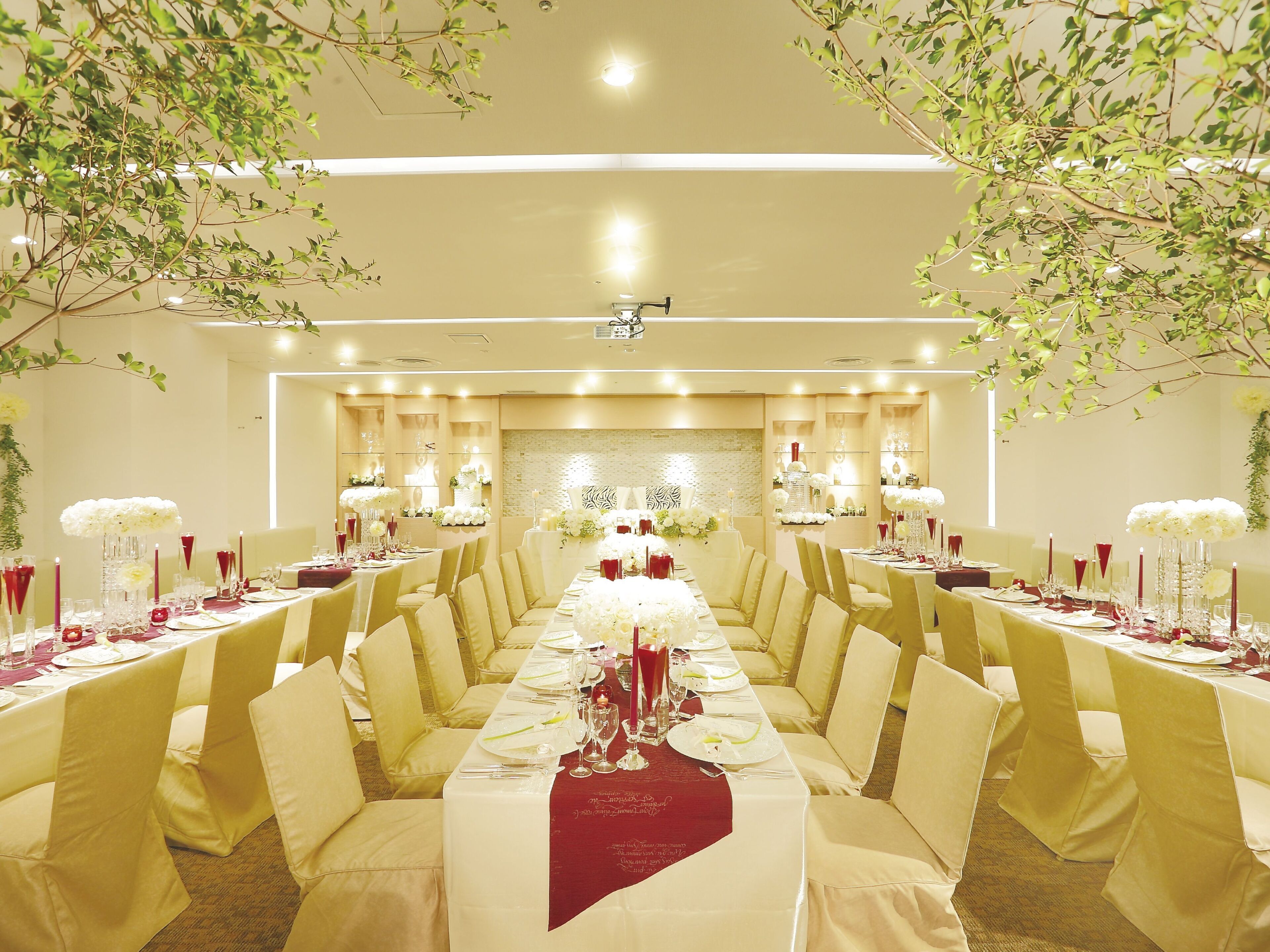 Un salón de banquetes con largas mesas dispuestas para un evento formal, con manteles blancos y rojos, velas y centros de mesa florales.
