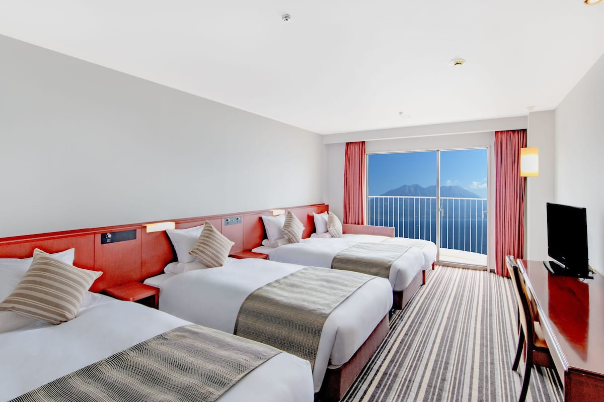 A hotel room with two beds, a TV, and a large window with a view of the sea.