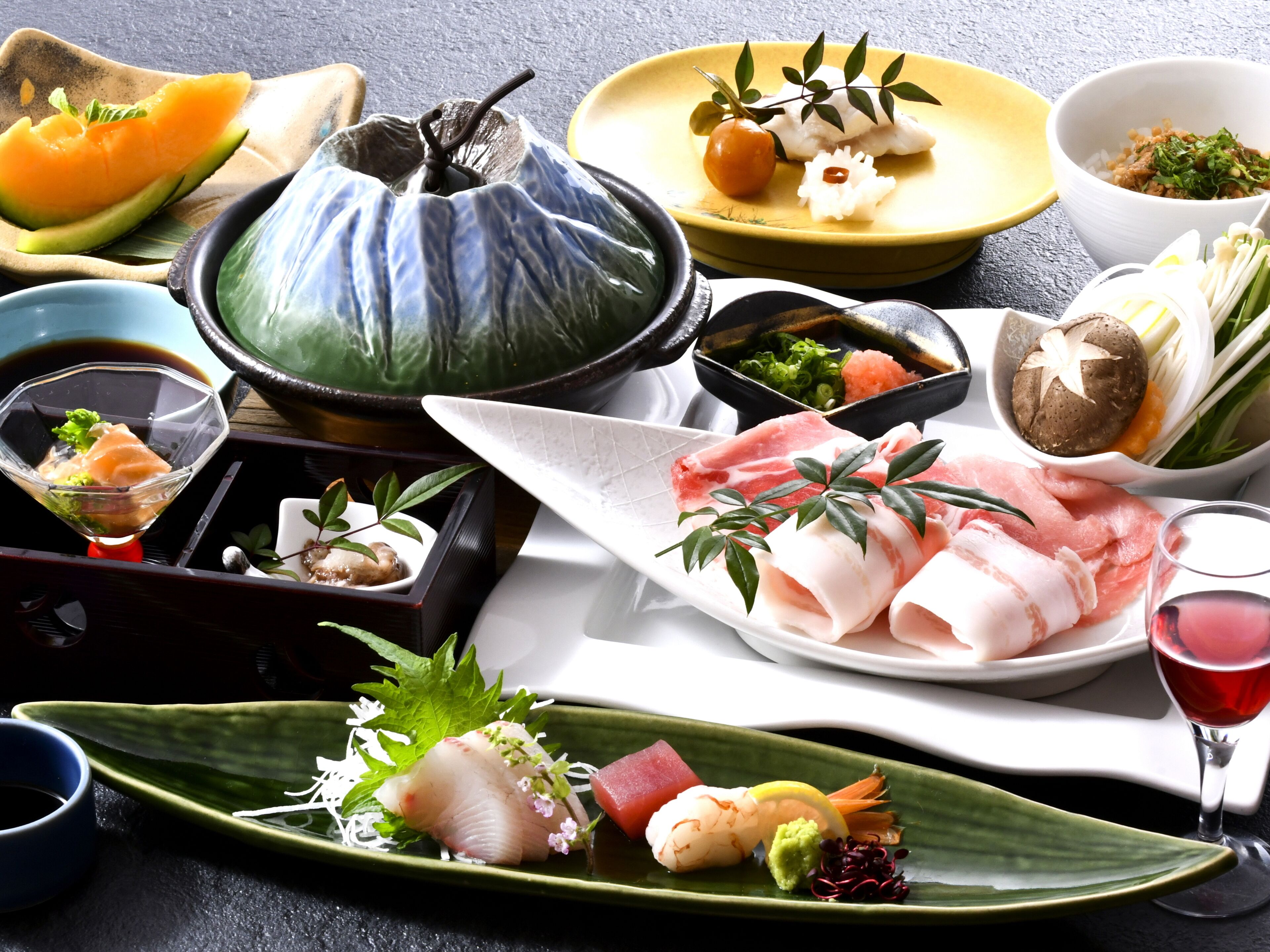 A variety of Japanese dishes, including sashimi, sushi, and tempura, arranged on a table with a teapot and a glass of red wine.