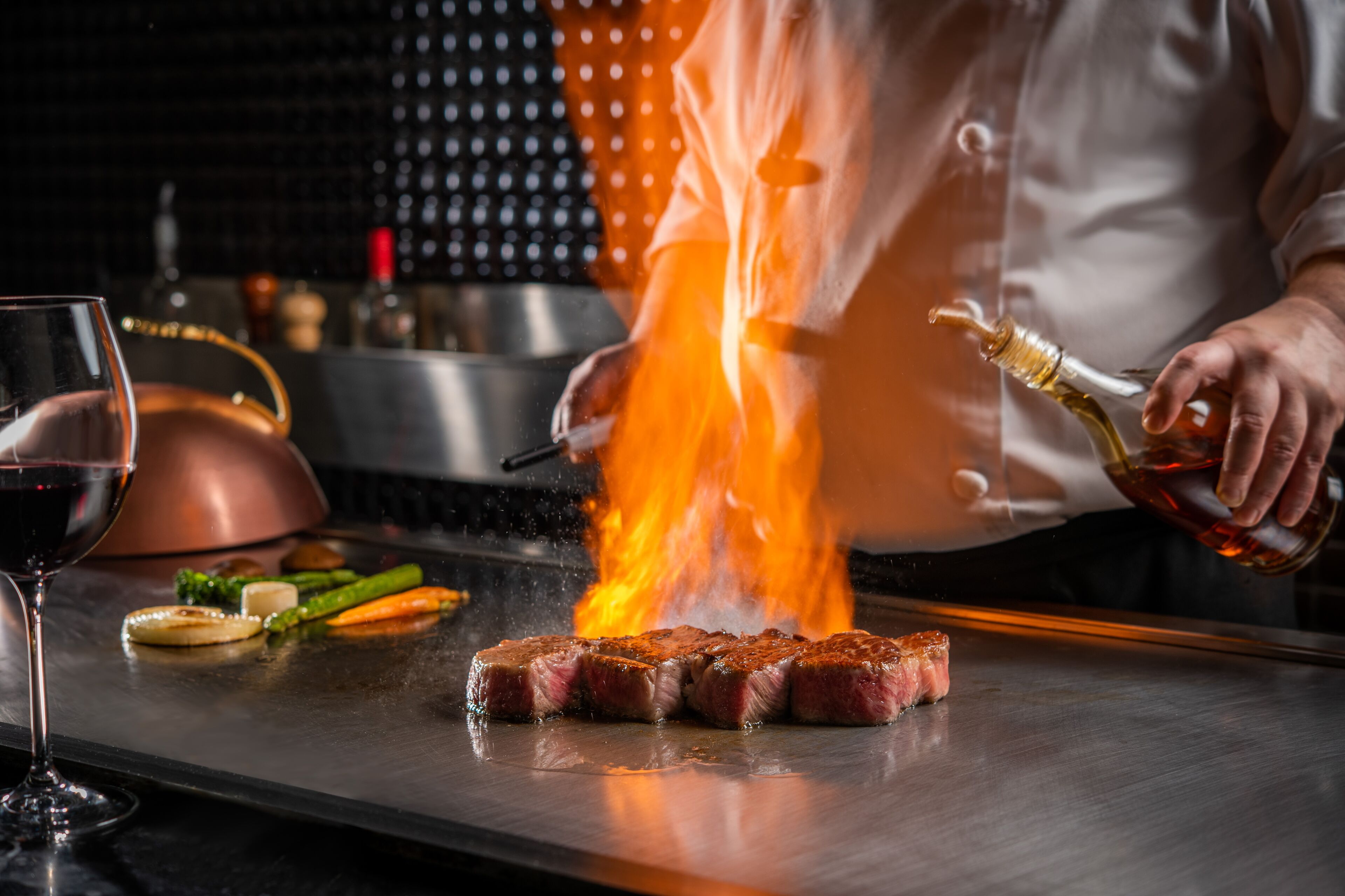 A chef cooking steak on a grill with flames, a glass of red wine, and vegetables on the side.