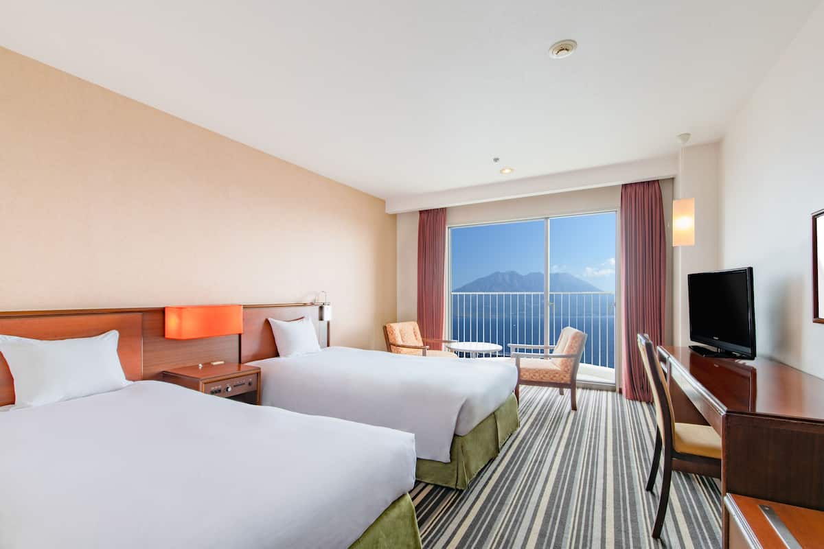 A hotel room with two beds, a TV, a desk, and a balcony with a view of the mountains.