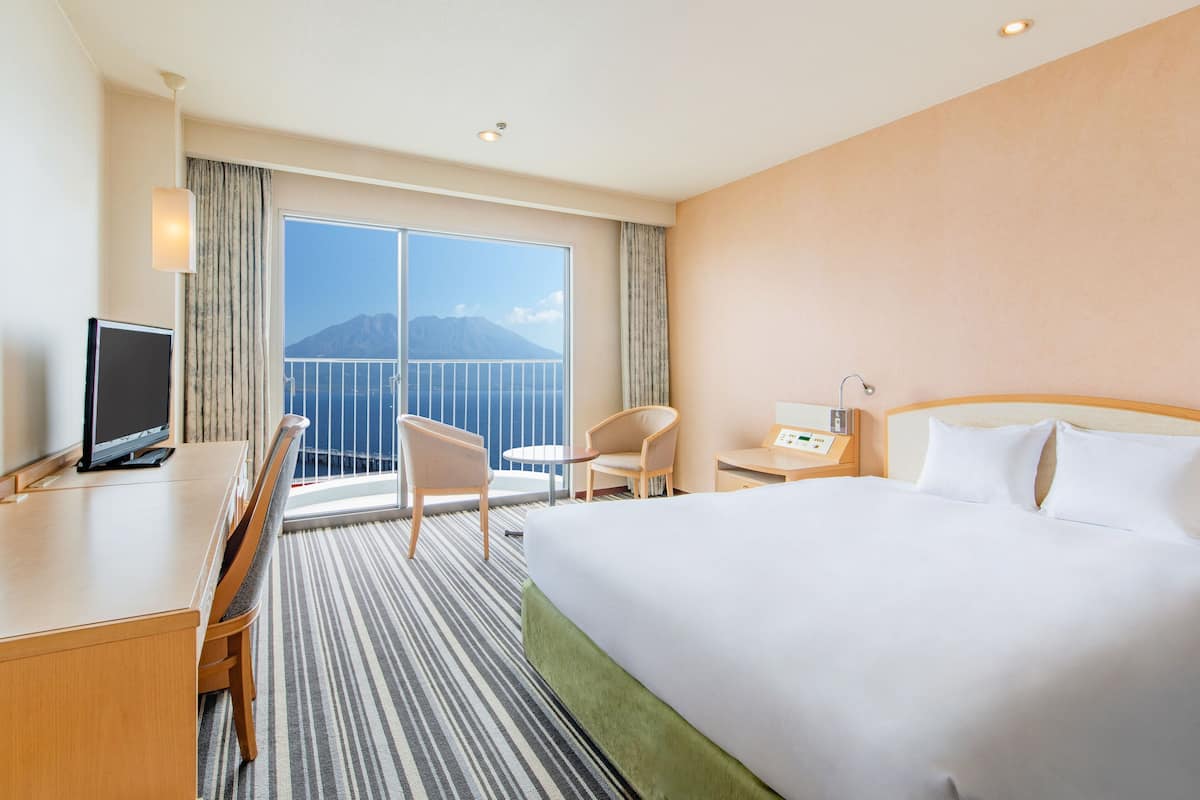 A hotel room with a bed, a desk, a chair, a TV, and a view of a mountain through a large window.