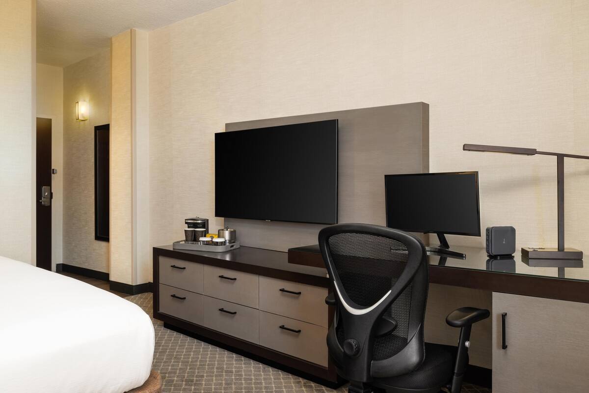 A hotel room with a bed, a desk with a computer, a chair, a flat-screen TV, and a lamp.