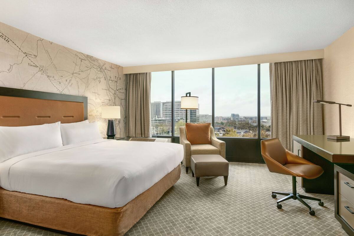 A hotel room with a large bed, a desk, a chair, and a view of the city through large windows.
