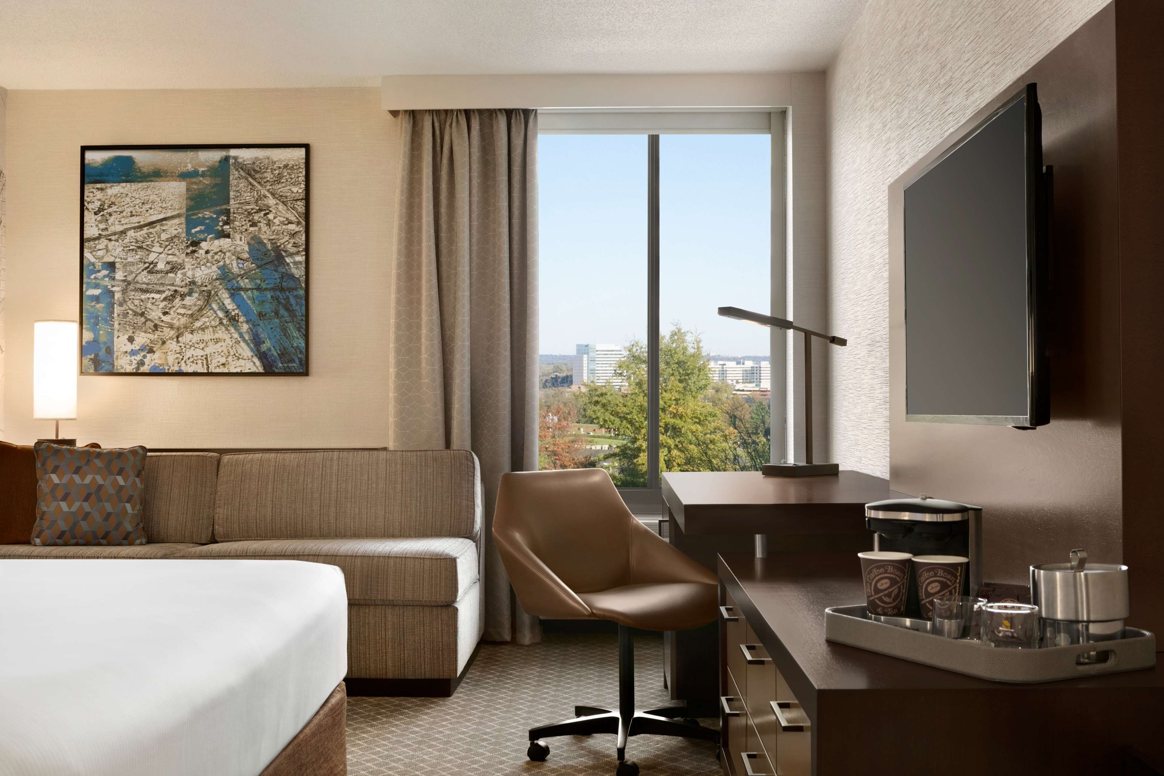 A hotel room with a bed, a sofa, a desk with a chair, a TV, and a window with a city view.