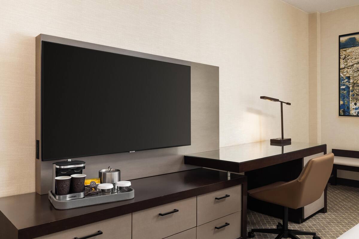 A hotel room with a large flat-screen TV, a desk with a coffee maker, a chair, and a tray with cups and condiments.