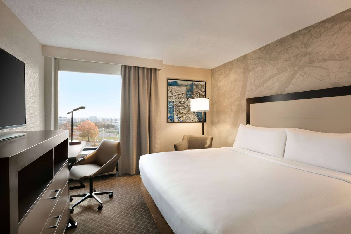 A hotel room with a large bed, a desk with a chair, a television, and a window with a view of buildings and trees.