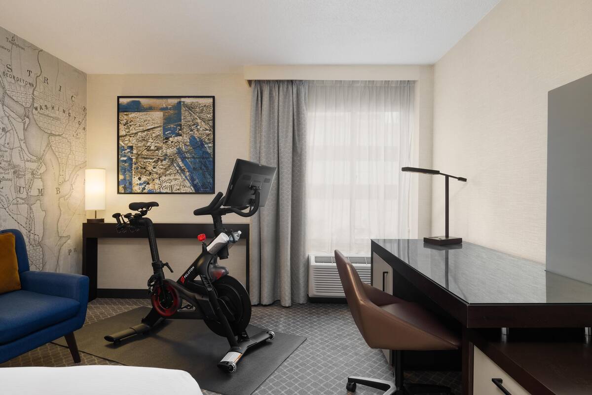 A hotel room with a desk, chair, exercise bike, map on the wall, and a large window with curtains.