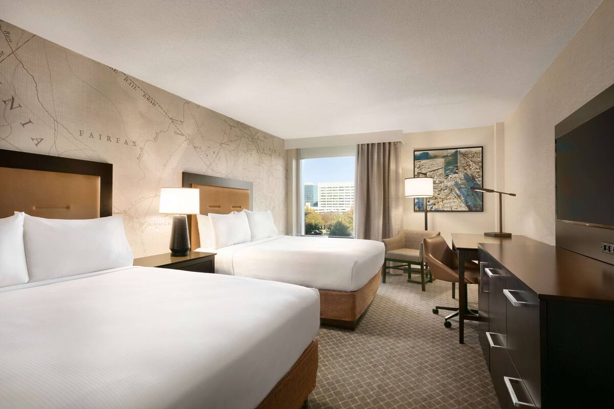 A hotel room with two beds, a desk, a chair, a TV, and a large window with a view of buildings.